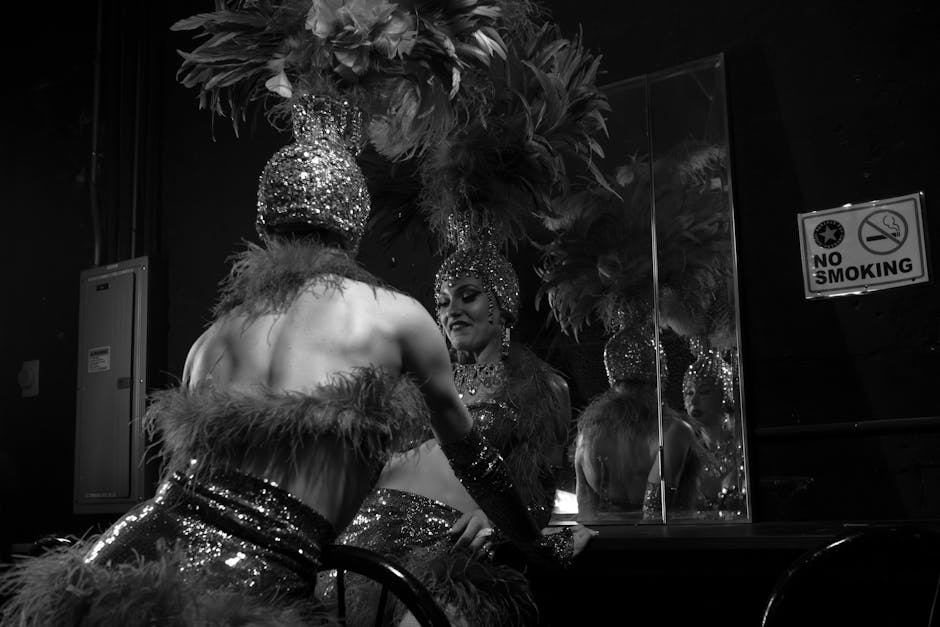 Dancer in feathered headdress and sequined costume gets ready backstage, reflecting in mirror.