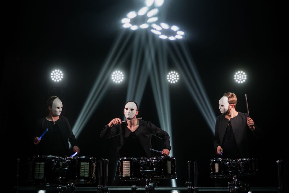 Three masked drummers perform energetically under dramatic stage lighting.